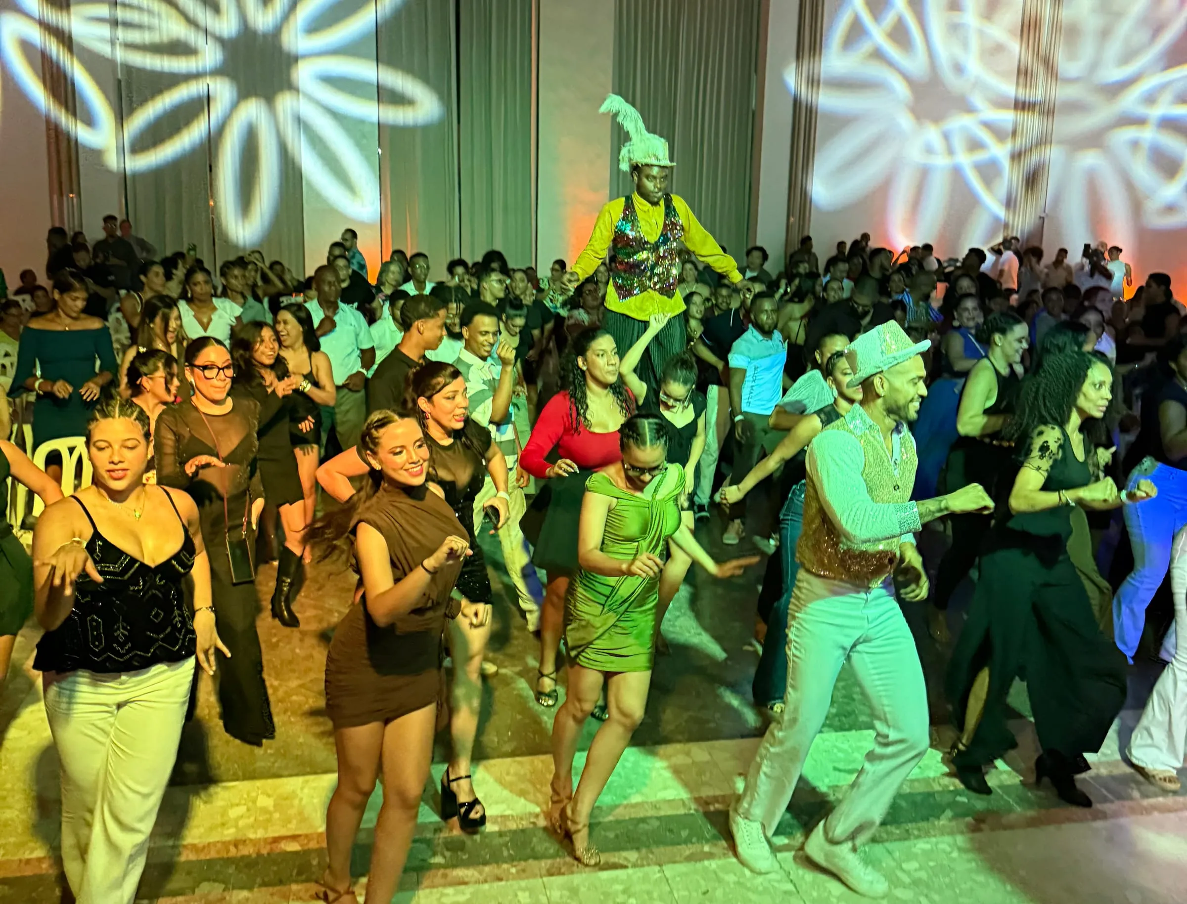 Social de Raíces: estudiantes y público bailando en un salón lleno, con un zanquero al fondo